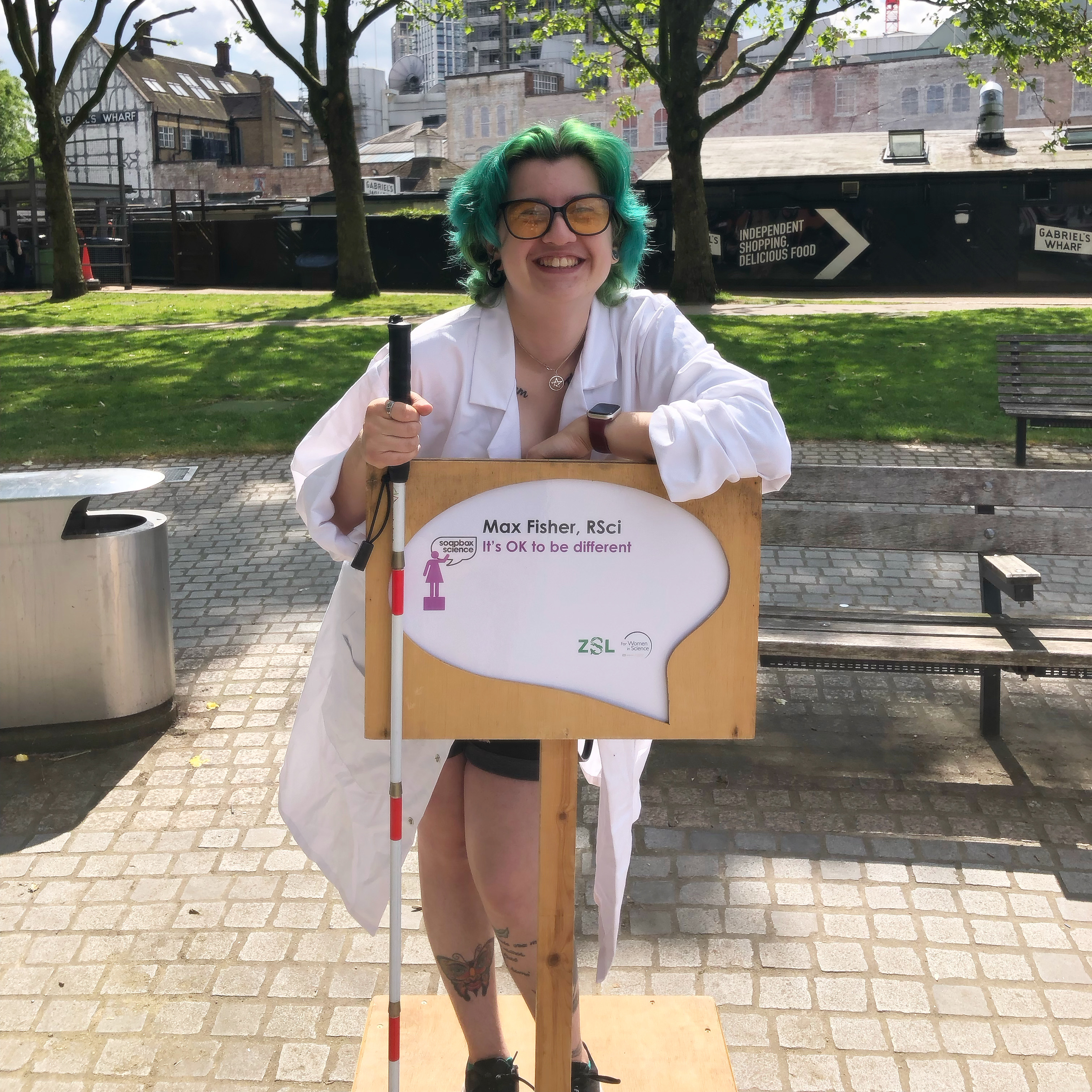 Max, a white nonbinary person with green hair, is wearing a lab coat while standing on a literal soapbox. They are holding their white cane and smiling. 