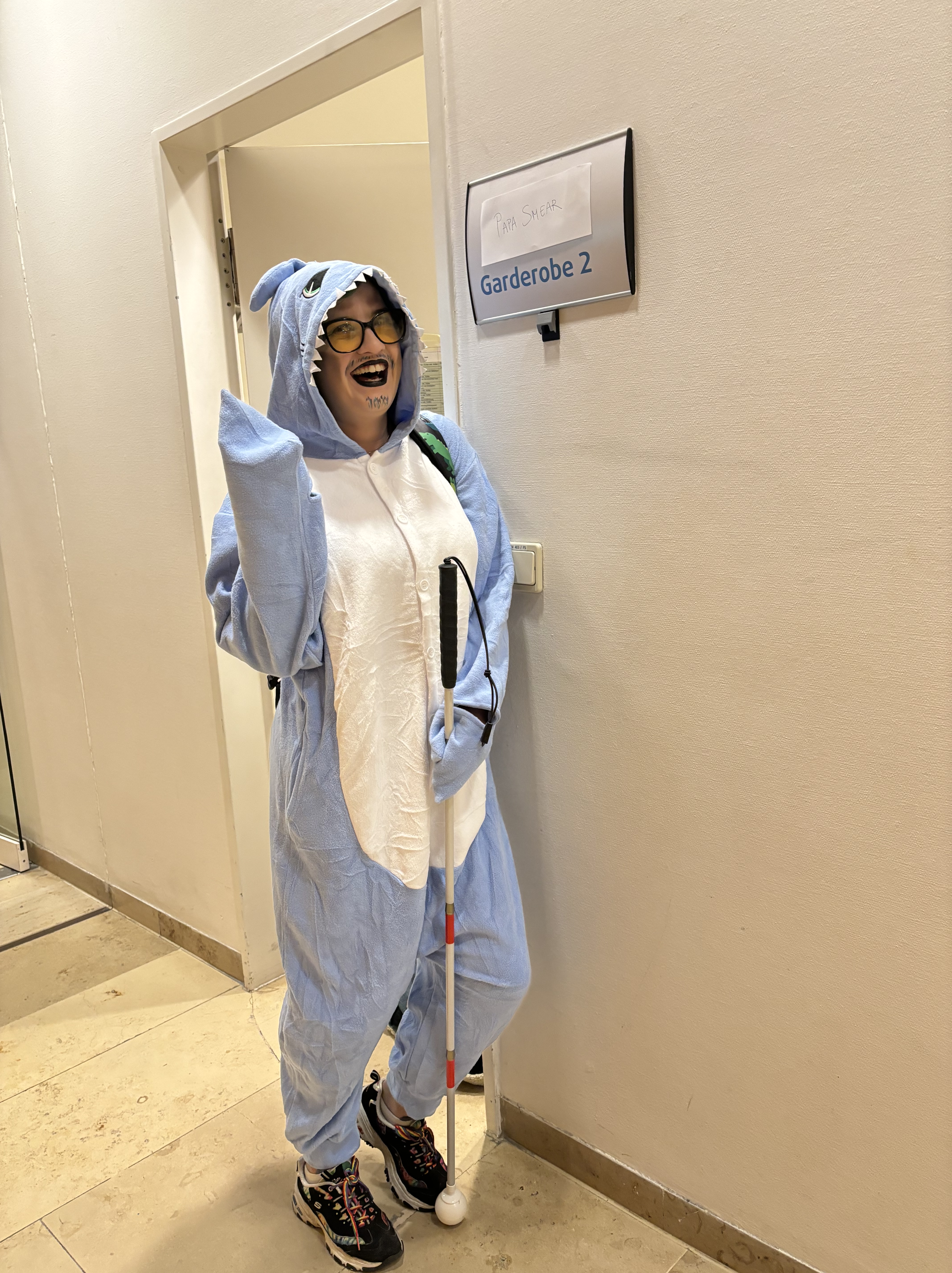 Max, a white nonbinary person, is dressing in a shark onesie in full drag, posing by their dressing room sign. 