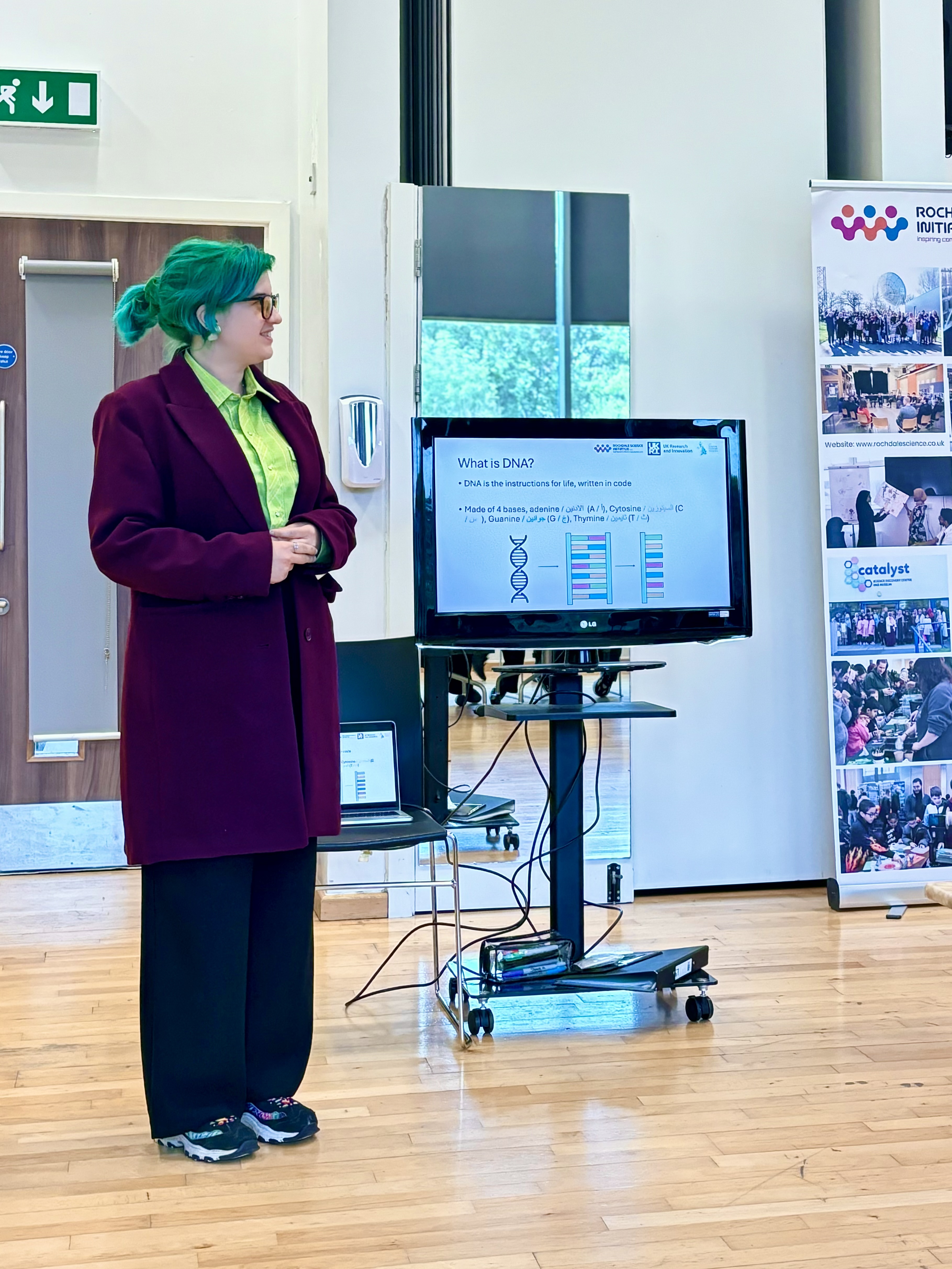 Max, a white nonbinary person, is standing at the front of a room teaching a group of Deaf adults about DNA transcription and translation.