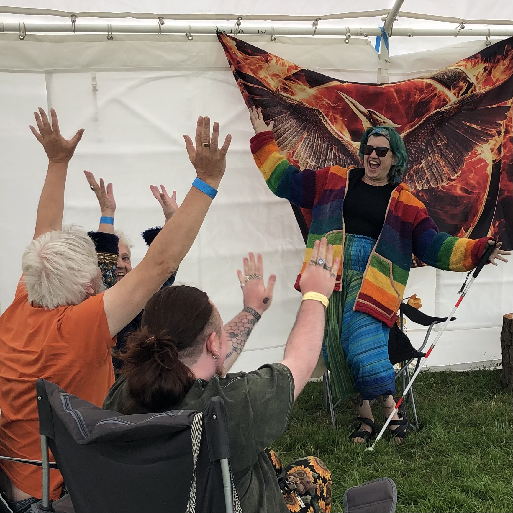 Max, a white nonbinary person with green hair is standing in a marque giving a talk on DeafBlindness in the pagan community. Max and the participants are waving their arms as if they are on a rollercoaster.