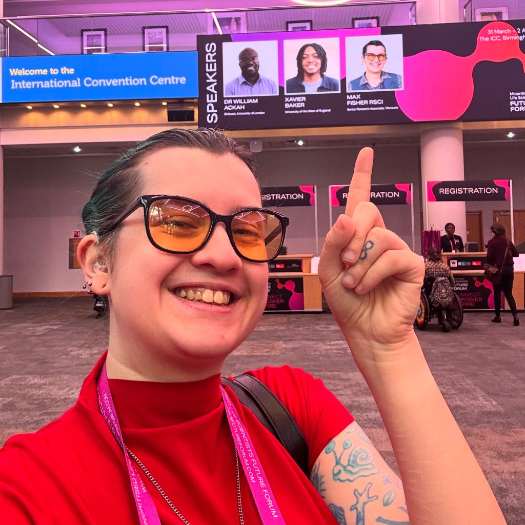 Max, a white genderqueer person with green hair, is taking a selfie and pointing at the billboard behind them with their face on it